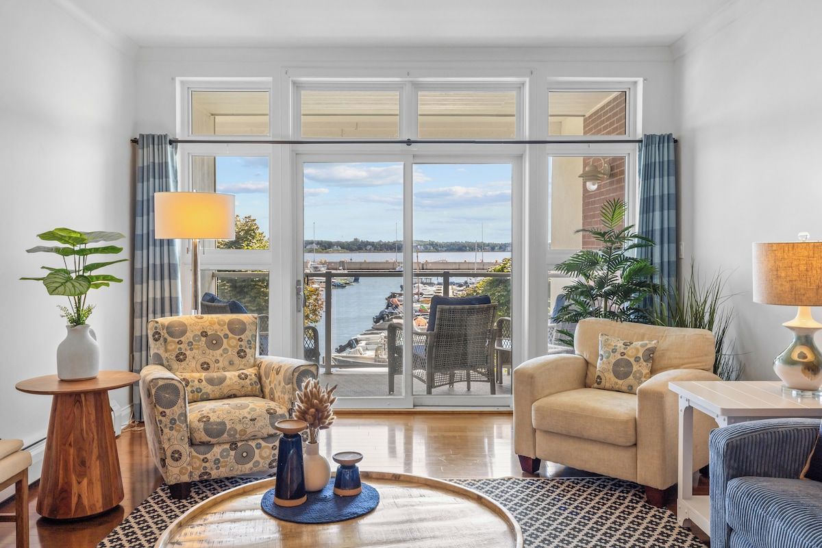 A spacious living room with a large sliding glass door leading to a balcony with a view of the water. There are two armchairs, a small table, and a lamp.