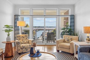 A spacious living room with a large sliding glass door leading to a balcony with a view of the water. There are two armchairs, a small table, and a lamp. - Luxury Waterfront Marina Condo in Downtown (Charlottetown)