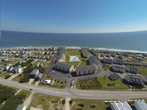 An aerial view of a coastal residential area with multiple apartment buildings, a swimming pool, and a road in the foreground.