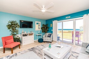 A room with a TV, a ceiling fan, a sliding glass door to the beach, a red chair, a white dresser, and a wicker table.