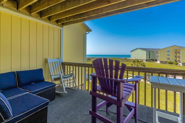A balcony with a view of the beach and ocean, a blue sofa, and two Adirondack chairs.