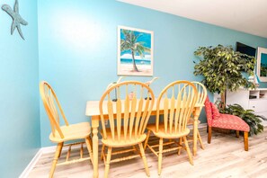 A dining area with a wooden table and chairs, a painting of a beach scene, and a potted plant.
