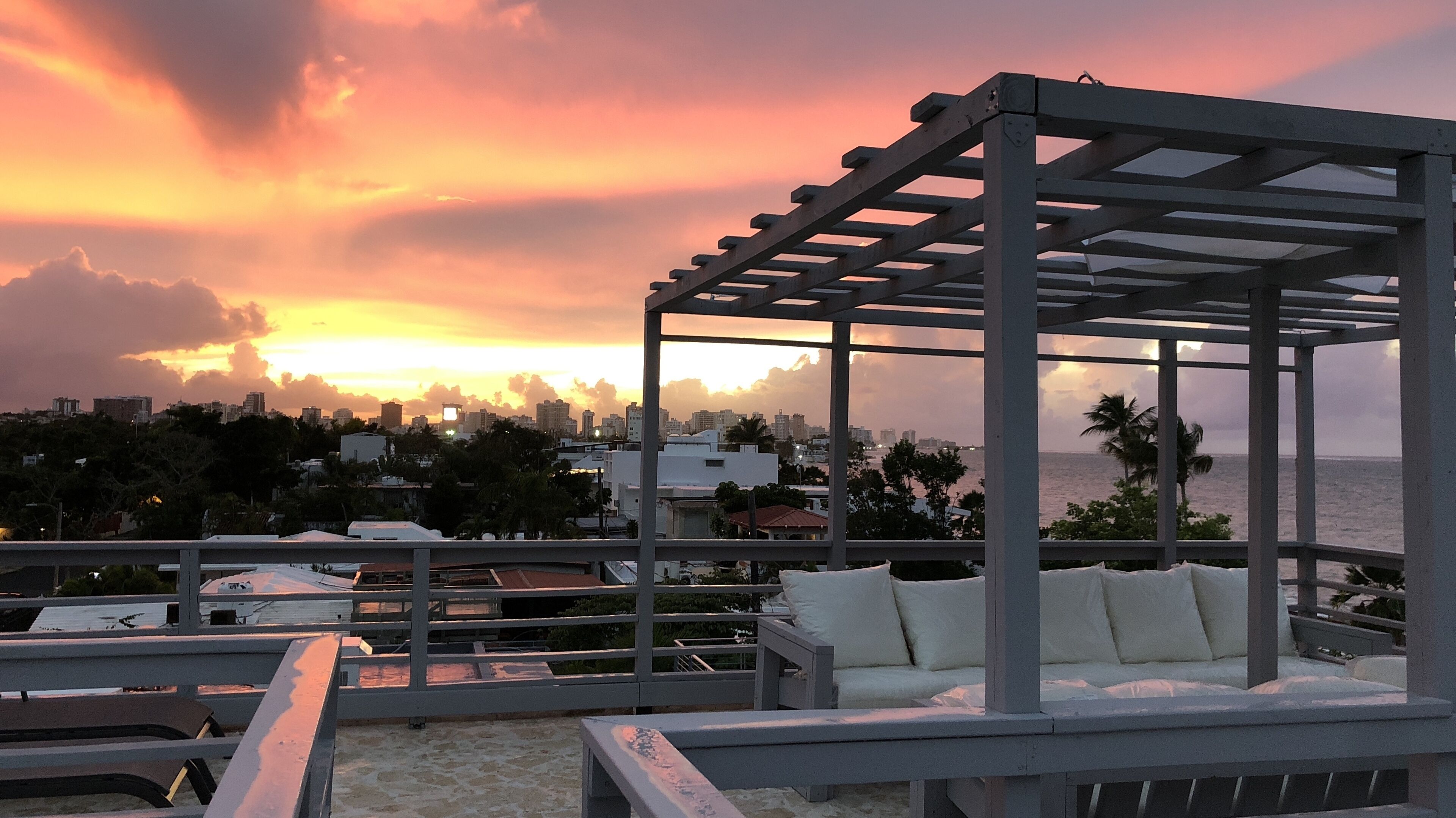 A rooftop terrace with a pergola and seating area, overlooking a city skyline and the ocean during sunset.