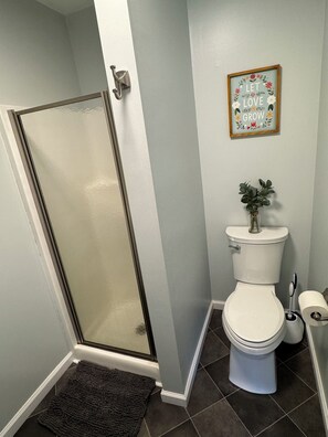 A bathroom with a glass shower door, a toilet, a small plant on the toilet tank, and a framed inspirational quote on the wall.