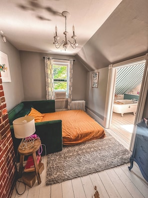Een slaapkamer met een bed, een radiator, een raam met gordijnen, een radiator zichtbaar door het raam, een houten vloer en uitzicht op een badkamer.