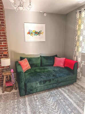 Une pièce avec un canapé vert, une lampe, un tableau encadré et un rideau à motifs.