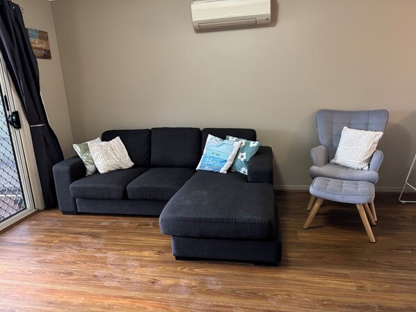 A modern living room with a dark grey sectional sofa, a grey armchair, and a small ottoman. A wooden floor and a wall-mounted air conditioning unit are visible. - Moonta Bay Family Getaway (Moonta Bay)