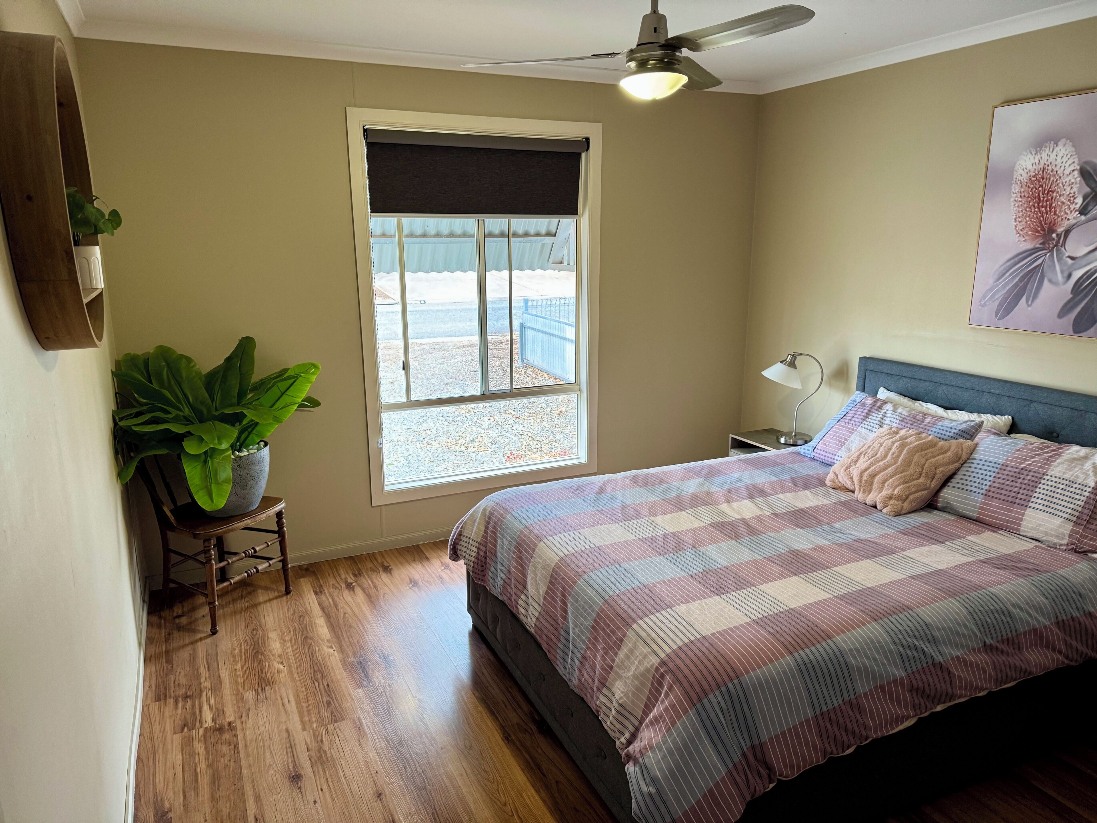 A bedroom with a bed, a ceiling fan, a window with a view, a bedside table with a lamp, and a potted plant.