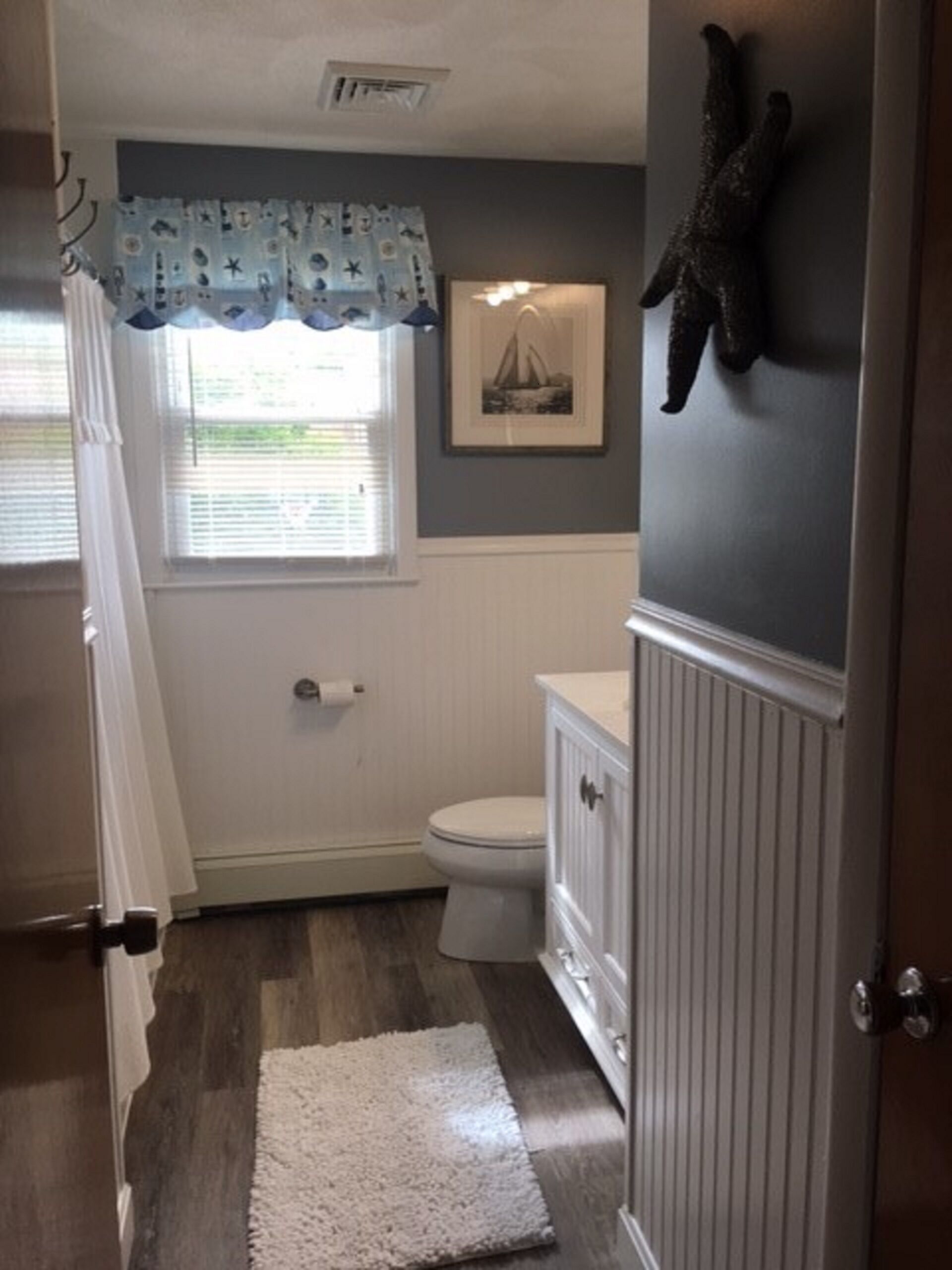 A bathroom with a toilet, a white vanity, a framed picture on the wall, and a window with a curtain.