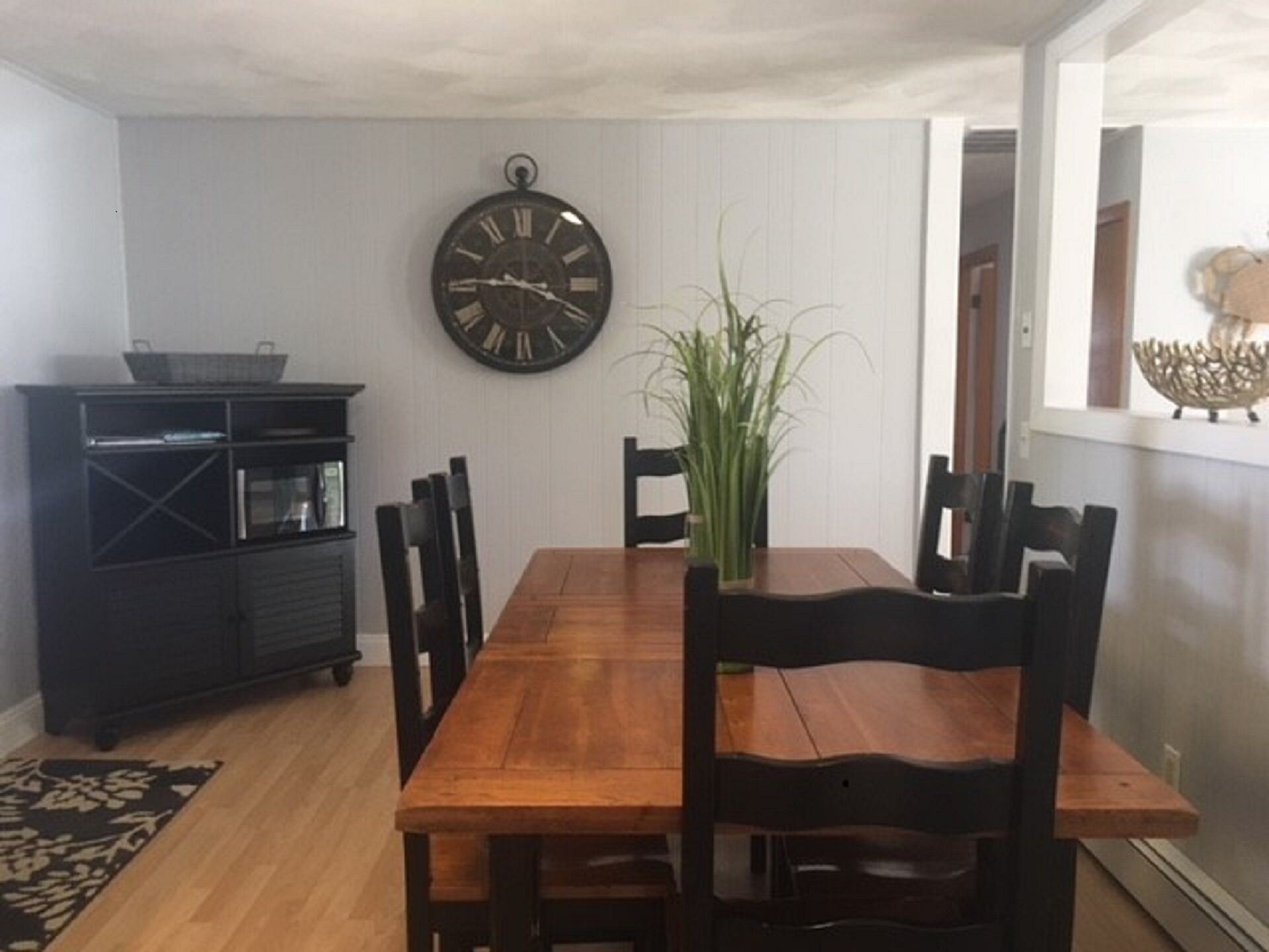 Een eethoek met een houten tafel, zwarte stoelen, een wandklok en een plant.