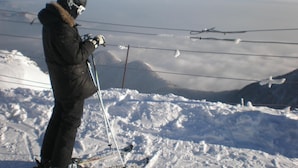 Une personne se tient debout sur une pente enneigée, équipée de skis, d’un casque et de gants, et tenant des bâtons de ski.