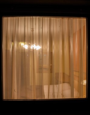 A room with a large window covered by sheer curtains, a bed, and a bedside table with a lamp.