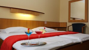 Une chambre d’hôtel avec un lit, deux boissons sur un plateau, un plaid rouge et une tête de lit en bois.
