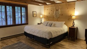 A bedroom with a large bed, wooden beams, a wooden headboard, two bedside lamps, and a window with blinds. - Lakefront Catskills Lodge (Monticello)