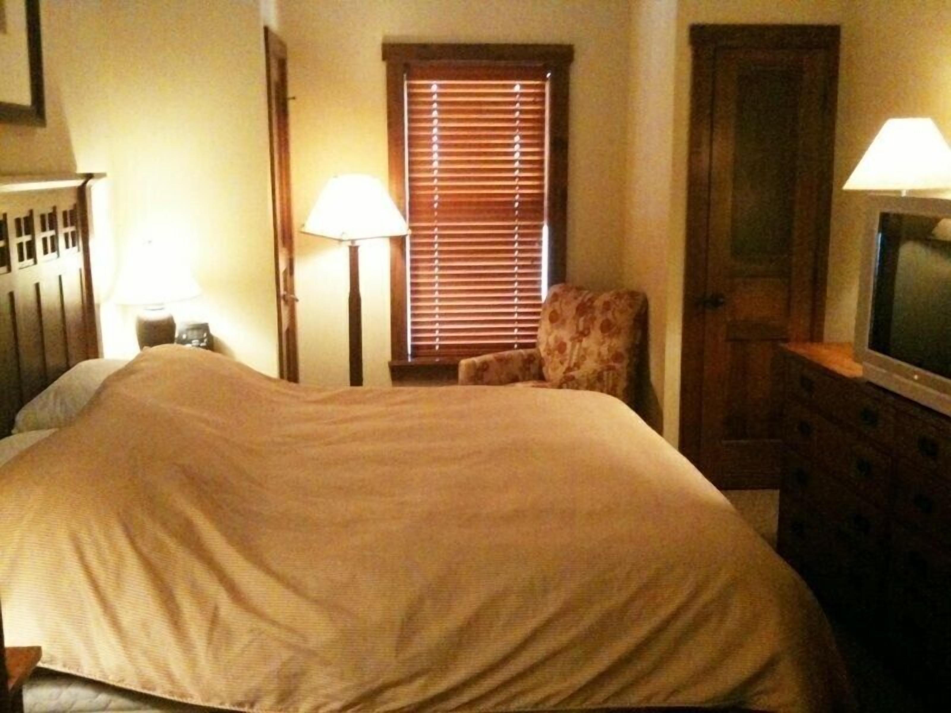 Une chambre d’hôtel avec un lit, une chaise, une commode, un téléviseur et une fenêtre avec des stores.