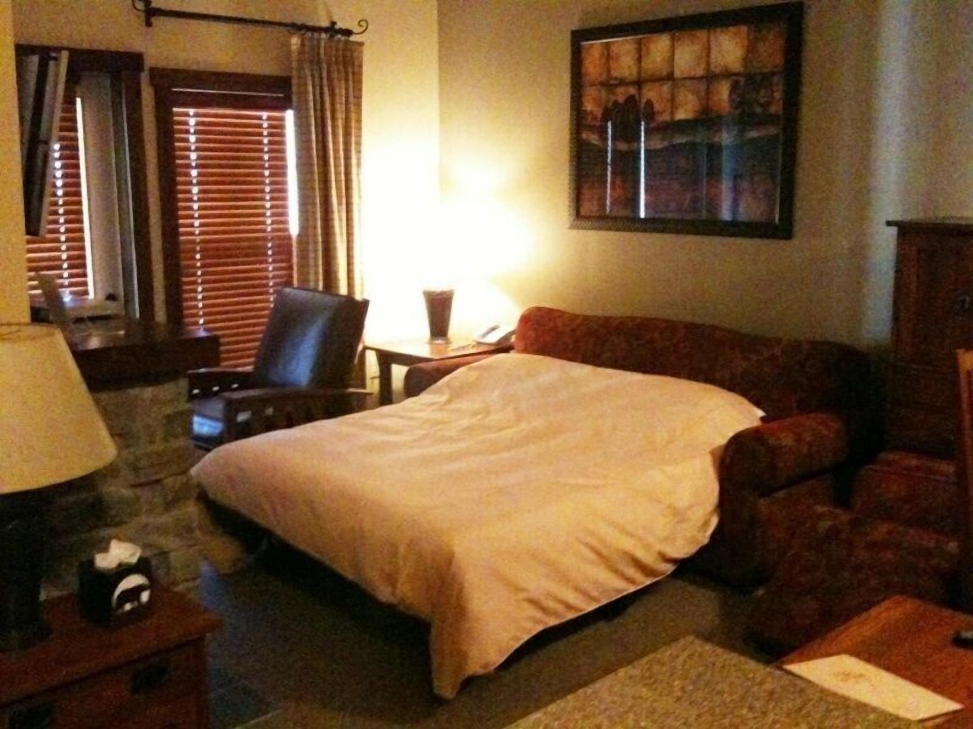 A hotel room with a bed, a chair, a desk, a lamp, and a painting on the wall.