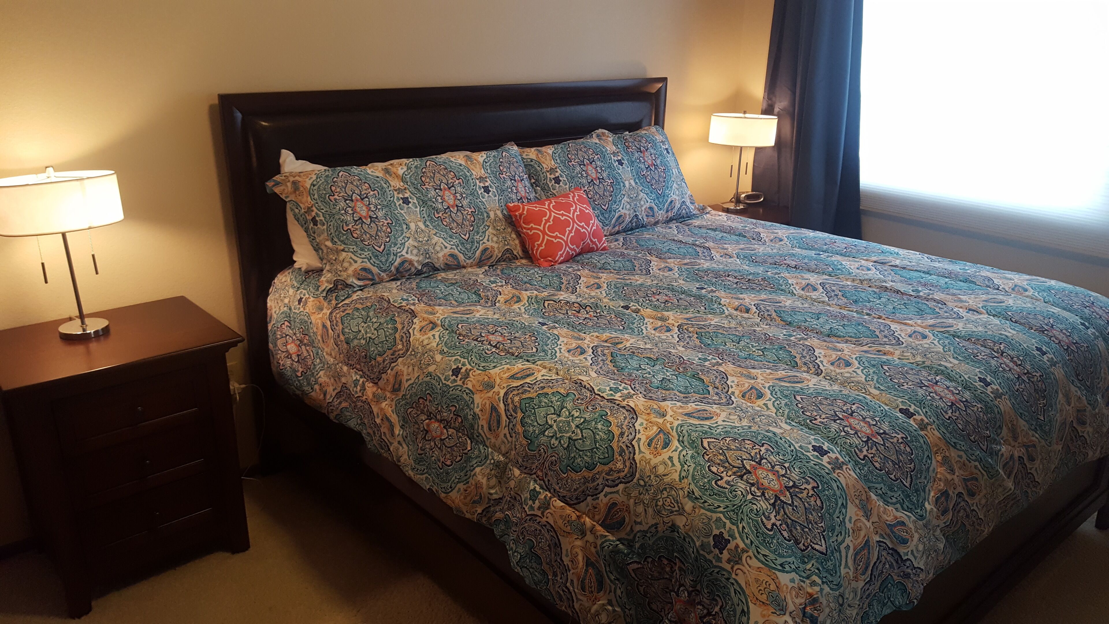 A neatly made bed with patterned bedding, two bedside tables with lamps, and a window with curtains.