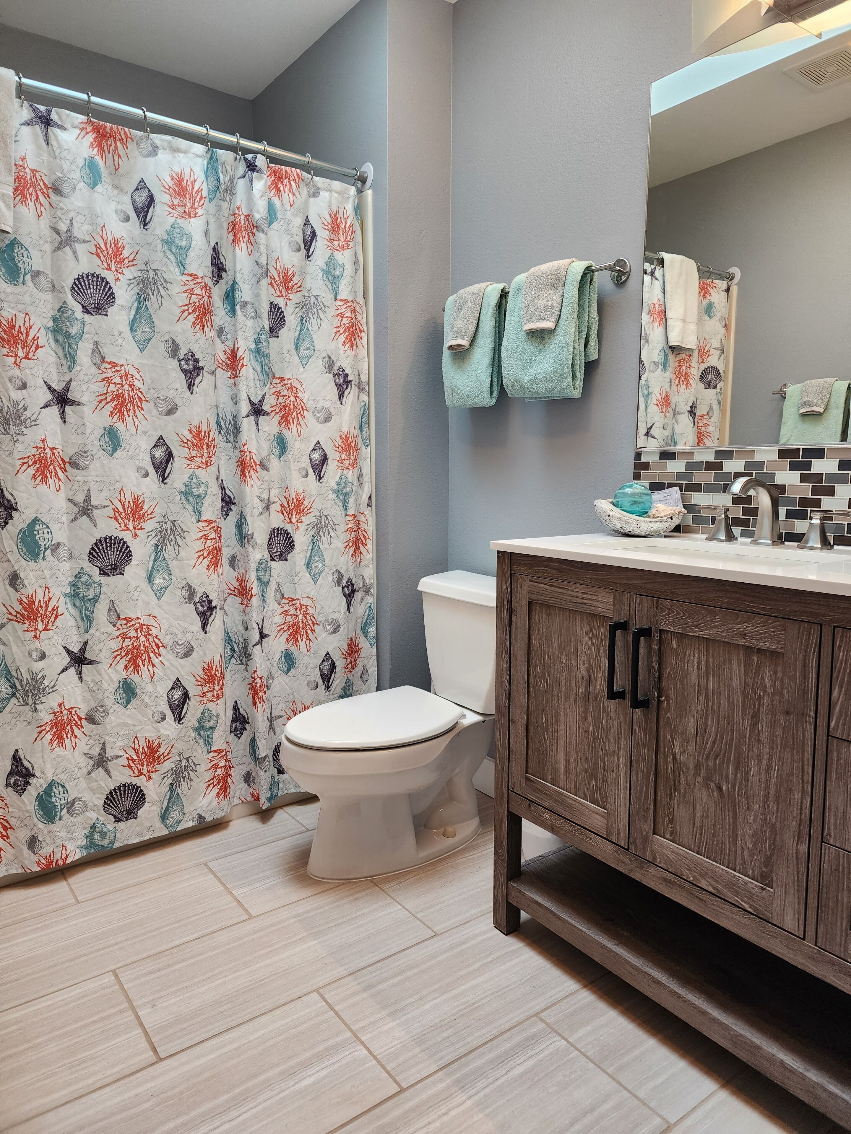 Une salle de bain équipée d’une douche avec un rideau orné d’un motif de coquillages, de toilettes, d’un meuble-lavabo en bois avec un miroir et d’un lavabo.