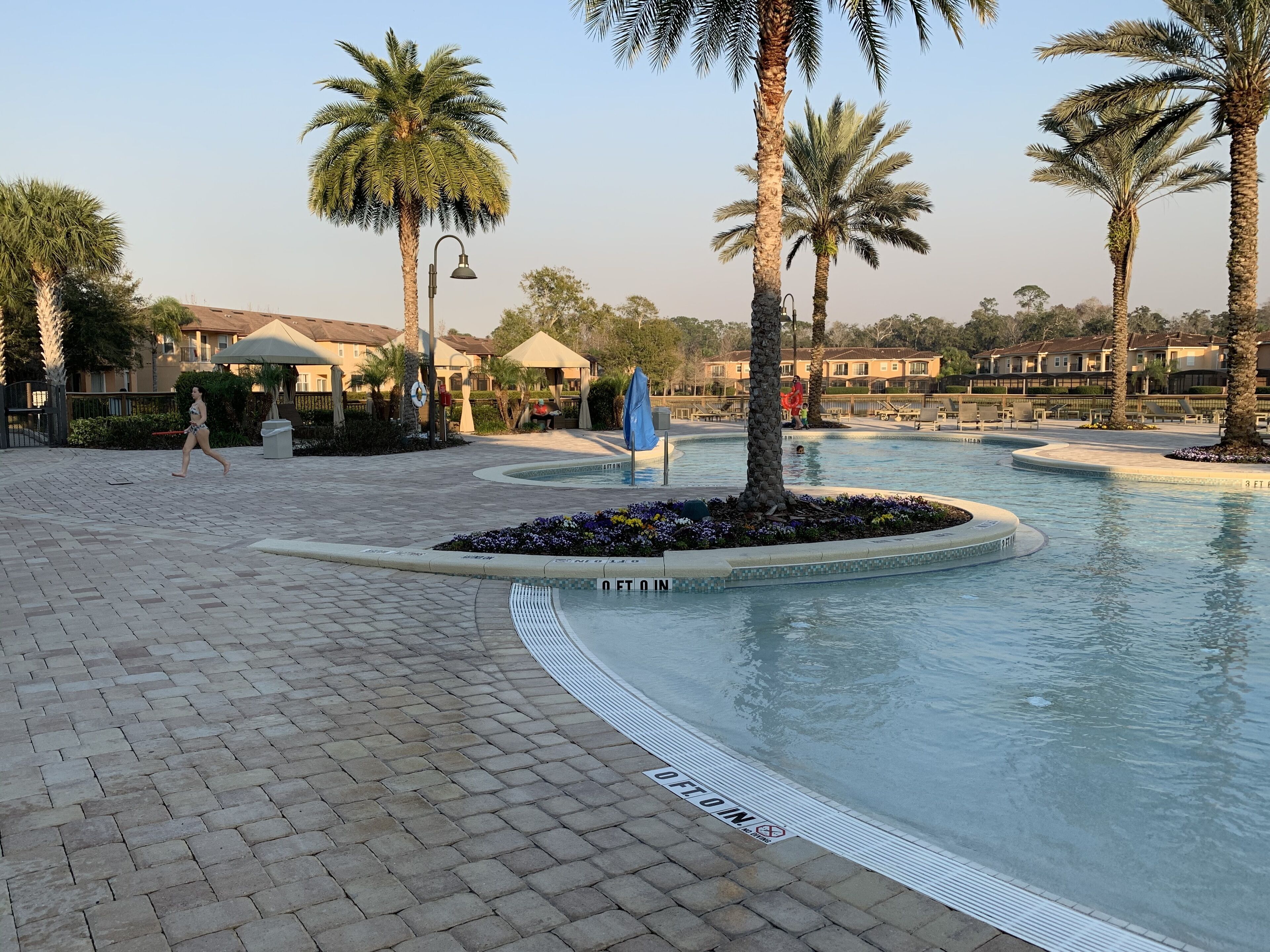 A swimming pool area with palm trees, a paved walkway, and a building with a roofed structure in the background.