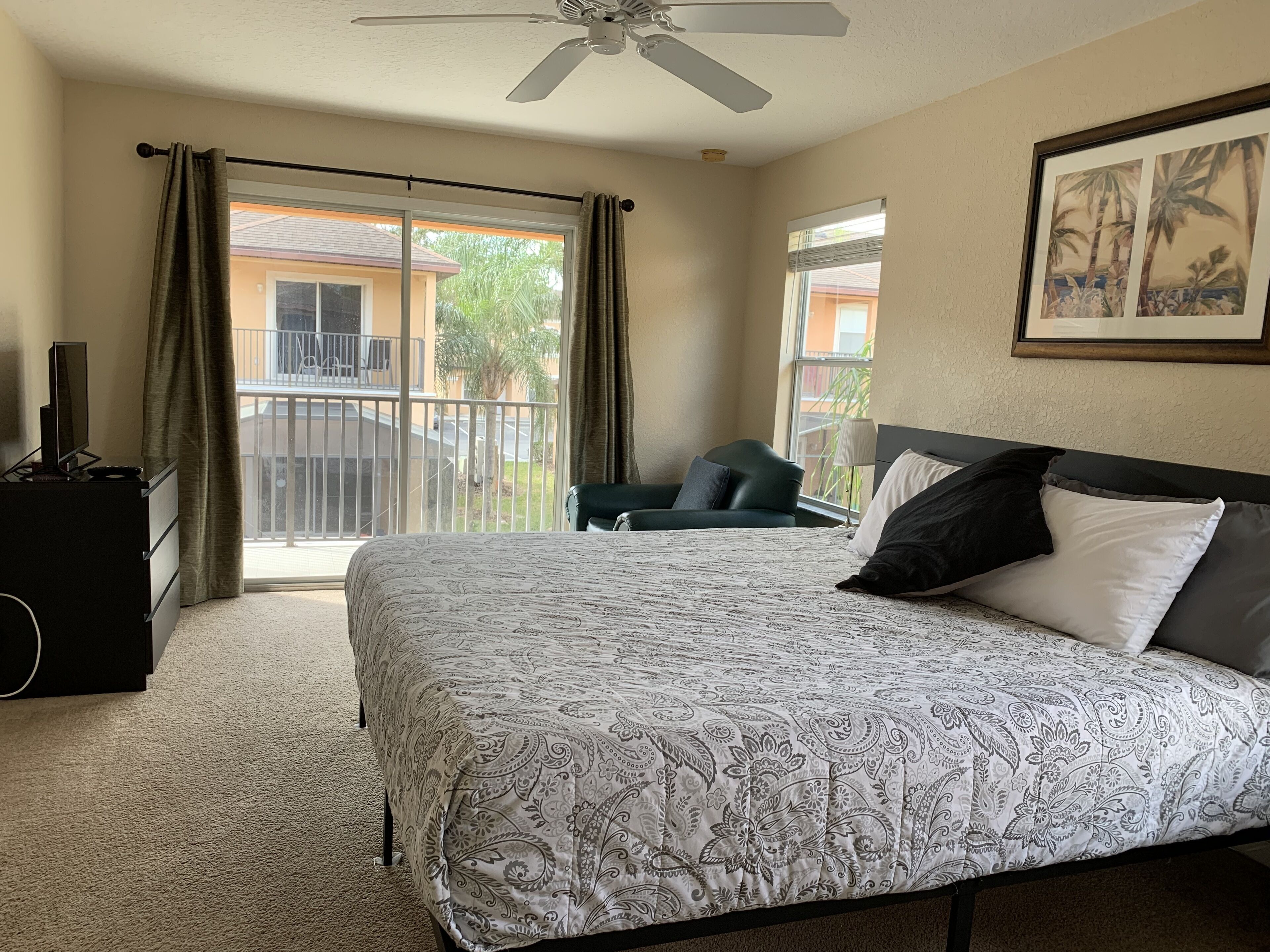 A hotel room with a large bed, a ceiling fan, a desk with a chair, and a view of the outside balcony.