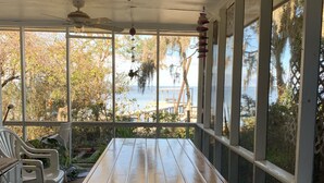 Un espacio al aire libre cubierto con mesa y sillas de madera, amplias ventanas que ofrecen vistas a la playa y árboles.