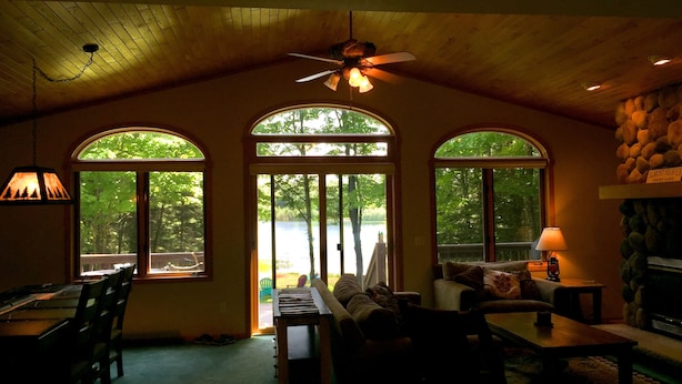 Une pièce chaleureuse avec un plafond en bois, un ventilateur de plafond et de grandes fenêtres en arc donnant sur un lac.