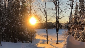 Un sentier enneigé mène à un banc d’où l’on peut admirer le soleil se coucher derrière les arbres.