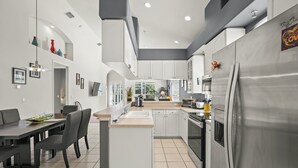 Eine moderne Küche mit weißen Schränken, einer zentralen Kochinsel, einem Essbereich mit Stühlen und einem großen Kühlschrank.