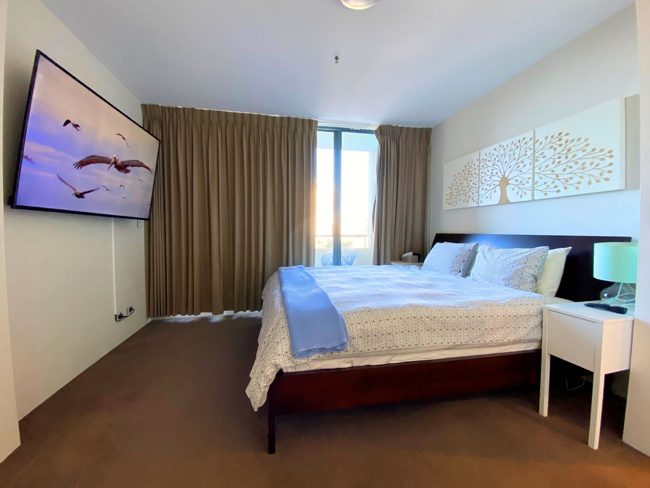 A hotel room with a large bed, a TV mounted on the wall, a window with curtains, and a wall art piece.