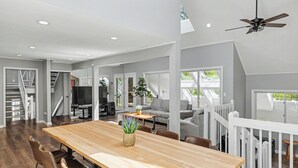 A spacious living area with a wooden dining table, comfortable seating, a television, and a staircase leading to an upper level. - Modern Creek-front House with Rooftop and Volleyball court (Bushkill)