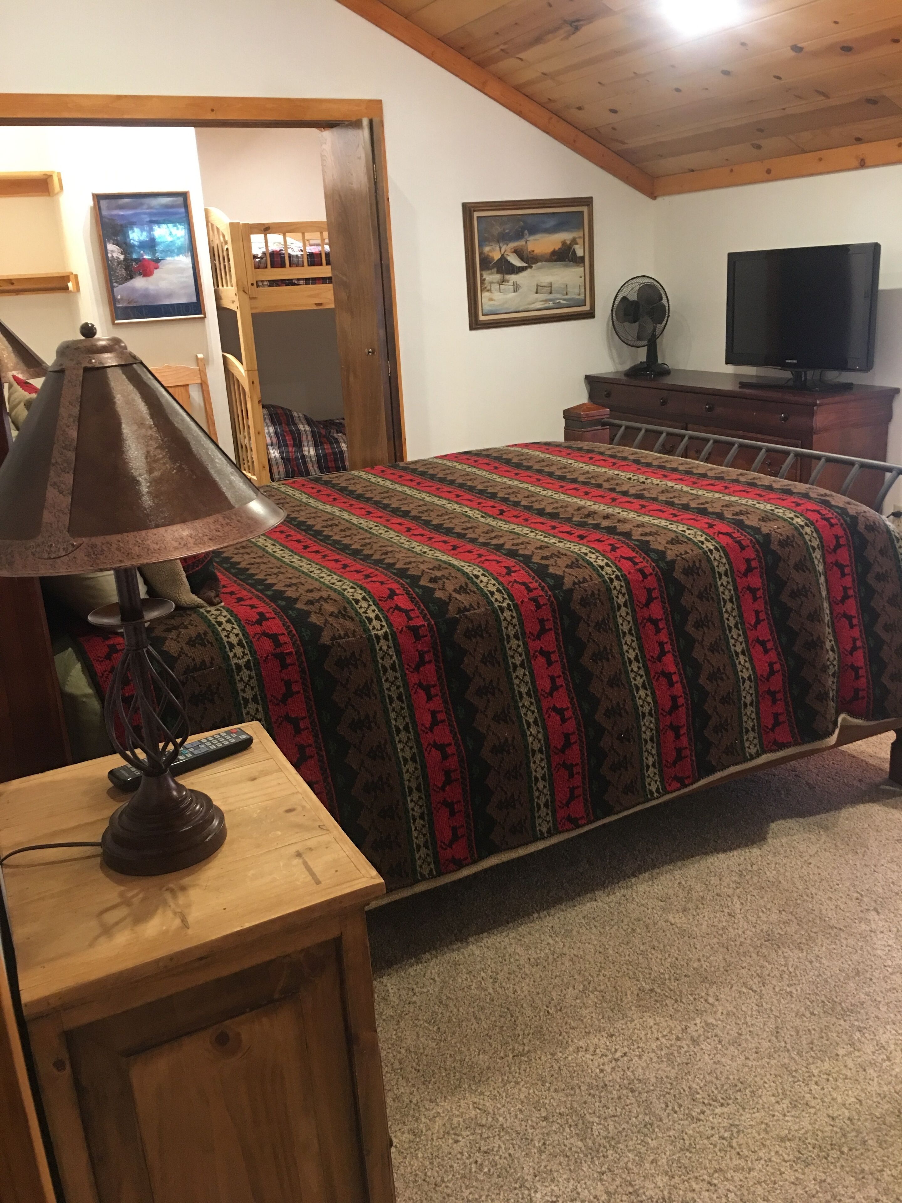 Een slaapkamer met een bed, een houten nachtkastje, een lamp, een televisie en een schilderij aan de muur.