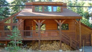A wooden cabin with a covered deck, a staircase leading to the main entrance, and a stack of firewood in front.