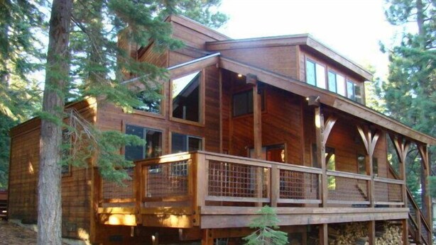 A wooden cabin with a deck, large windows, and a staircase leading to the upper level.