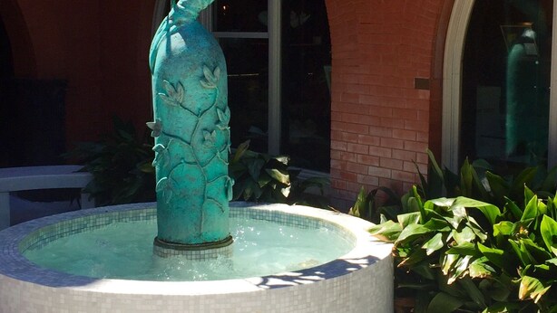 A bronze statue of a woman holding an umbrella, standing in a fountain with a tiled pool, set against a red brick building with arched windows.