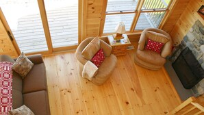 A cozy cabin interior with a wooden floor, a sofa, two armchairs with patterned cushions, a small table with a lamp, and a fireplace. - Elegant log home with unobstructed ocean views (Cutler)