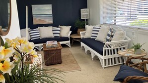 A cozy living room with a dark blue accent wall, a white wicker sofa set, a small round table, and a large window with blinds.