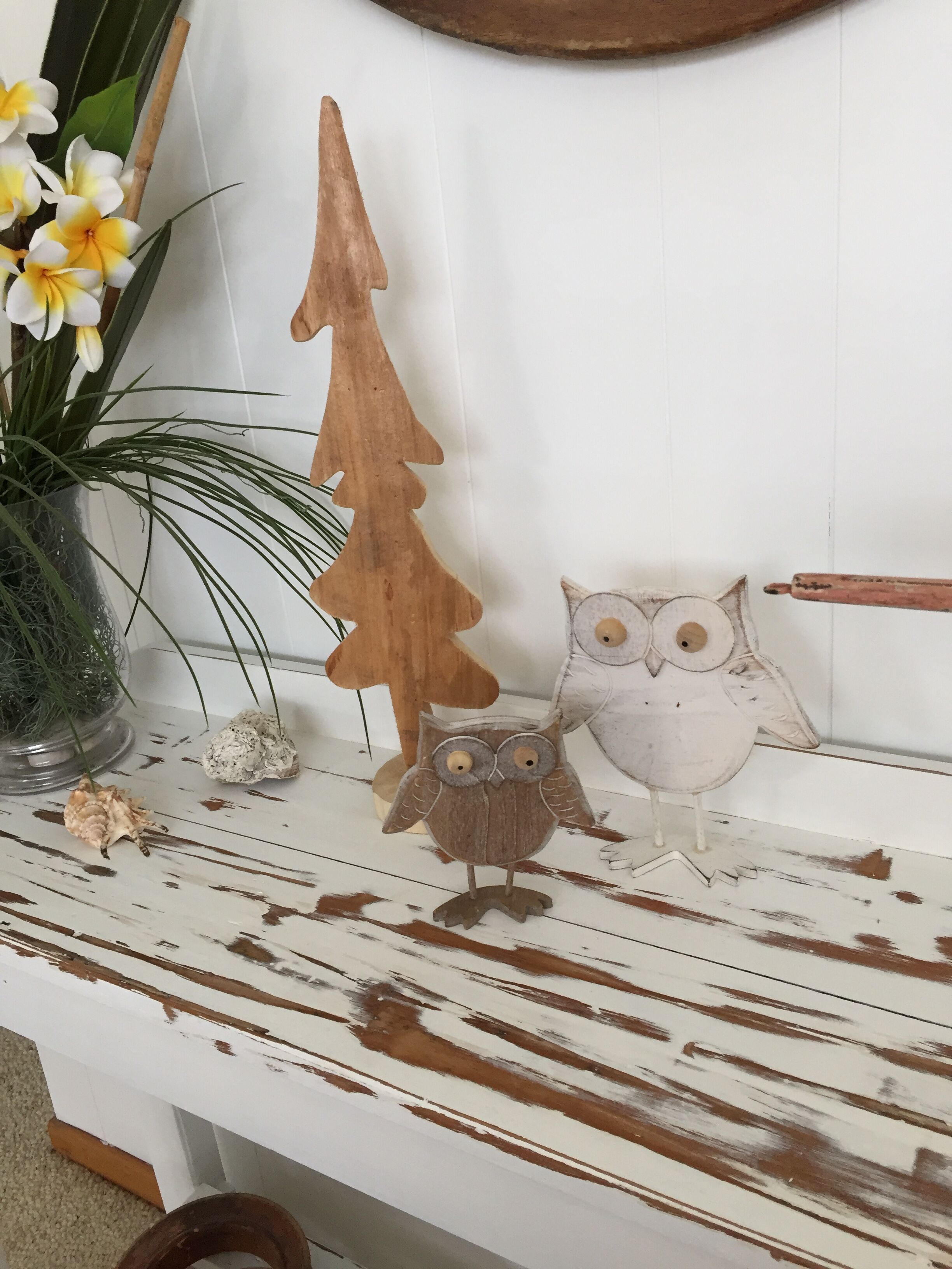 Een houten plank met een rustieke uitstraling, waarop een houten kerstboom, twee uiltjes en een bloempot met gele bloemen staan.