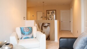A living room with a white sofa, a glass coffee table, and a blue patterned pillow. There is a painting on the wall and a door in the background.