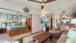 Ein geräumiges Wohnzimmer mit einer Couch, einem Couchtisch und einem Fernseher. Es gibt einen Deckenventilator, einen Wandfernseher und eine Zierwandverkleidung.