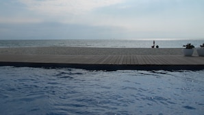 Une promenade en bois mène à une plage de sable fin, bordée par une mer calme et surplombée d’un ciel nuageux.