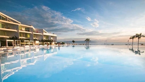 Une piscine d’un complexe hôtelier, avec une eau d’un bleu limpide, entourée de bâtiments modernes et de palmiers.