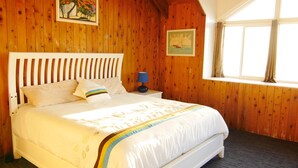 A bedroom with a wooden paneled wall, a bed with white bedding, a nightstand with a lamp, and a window with curtains. - Las Breezes 5 Bedroom Villa, With 1/2 Mile Private Beach On Hooper's Bay (George Town)