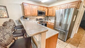 A kitchen with wooden cabinets, a stainless steel refrigerator, a microwave, and a stove. There is a bar area with stools. - 2 Night MIN Only MDW | 2 Pools/4 Hot Tubs (New Braunfels)