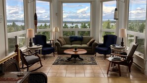 A spacious living room with large windows offering a scenic view of a lake and forest. There are comfortable seating areas, a coffee table, and a fireplace. - Island House Above Trees! 360° Oceanview, 4BR, Car-Accessible, Near Lunenburg   (La Have Island)