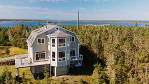 A multi-story house with a balcony, surrounded by dense forest and a body of water in the background. - Island House Above Trees! 360° Oceanview, 4BR, Car-Accessible, Near Lunenburg   (La Have Island)