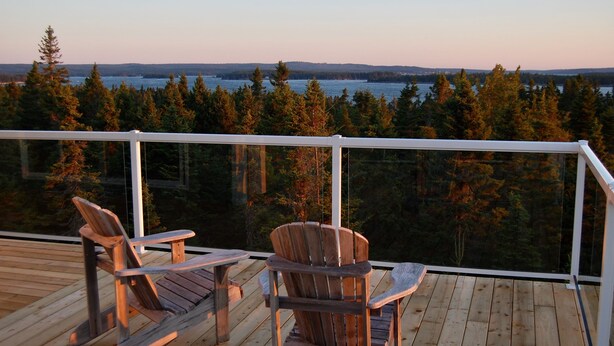A wooden deck with two Adirondack chairs and a table, overlooking a forested area and a body of water. - Island House Above Trees! 360° Oceanview, 4BR, Car-Accessible, Near Lunenburg   (La Have Island)