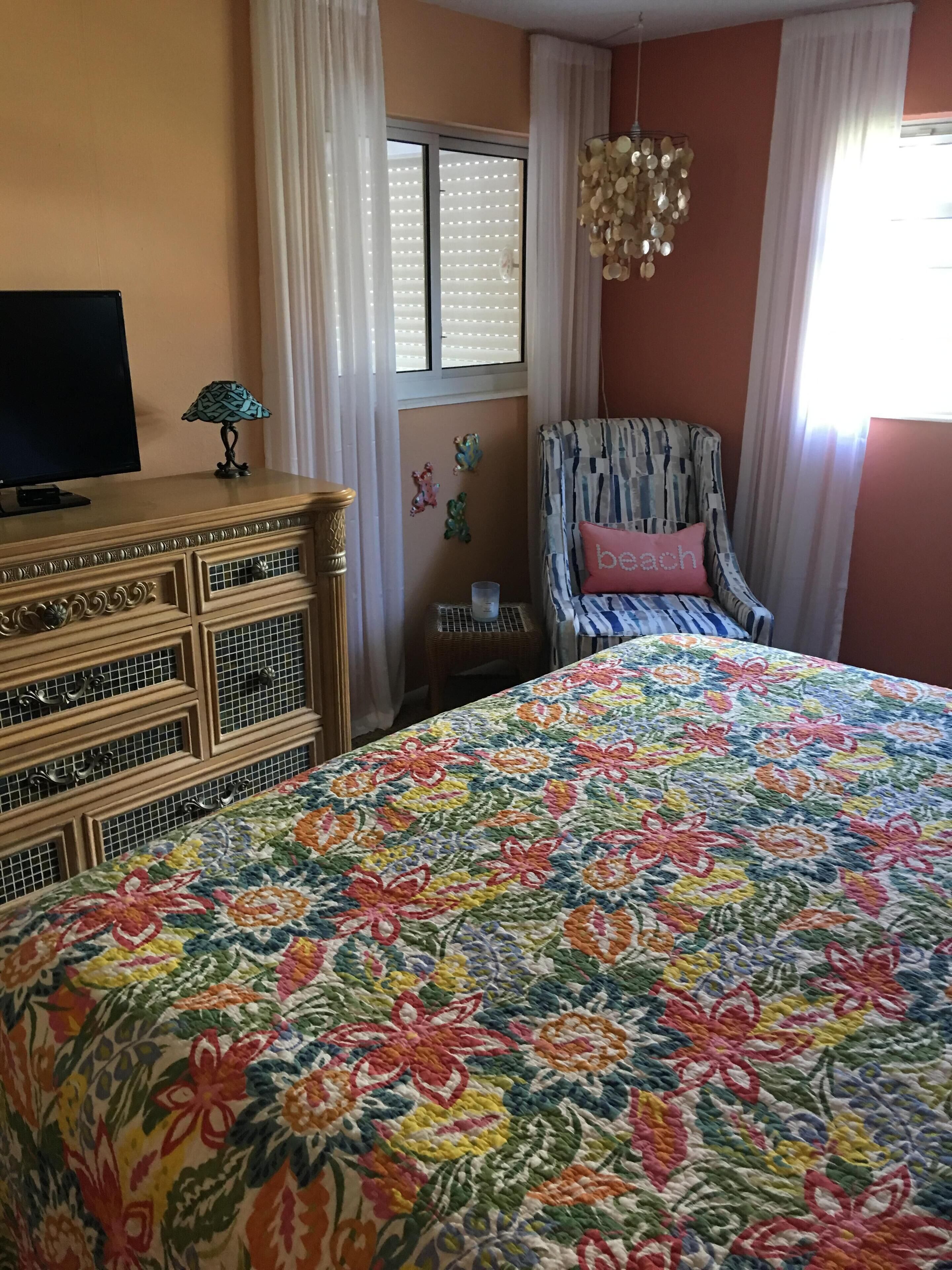 Une chambre à coucher avec une couvre-lit à motifs floraux, une commode en bois, un téléviseur, un fauteuil à motifs et une fenêtre avec des stores.