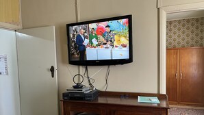 Un téléviseur fixé au mur affiche une émission, un meuble en bois se trouve en dessous, contenant divers objets, et une porte est située à gauche.