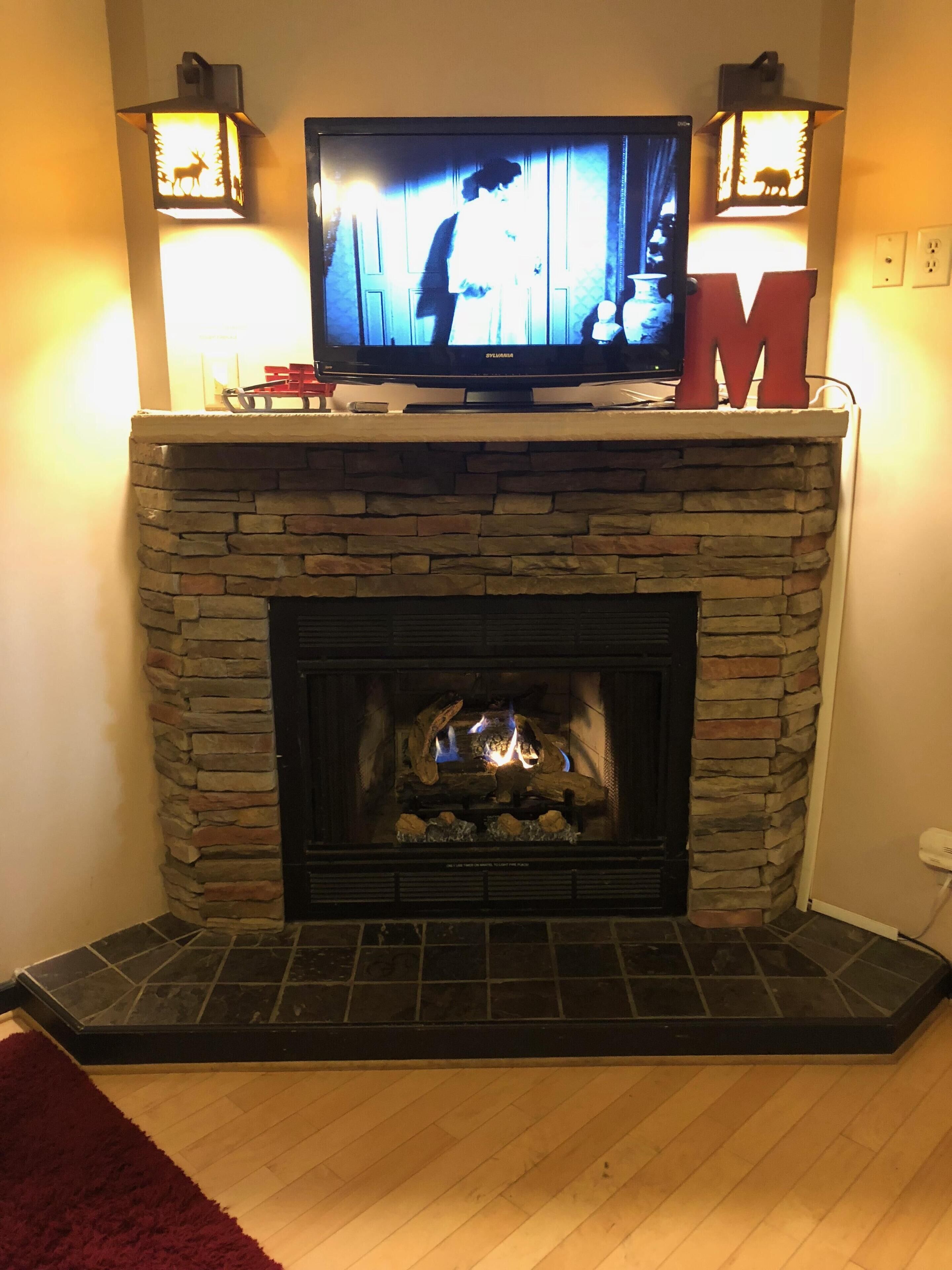 A fireplace with a television above it, a red 'M' decoration, and two wall-mounted lanterns.