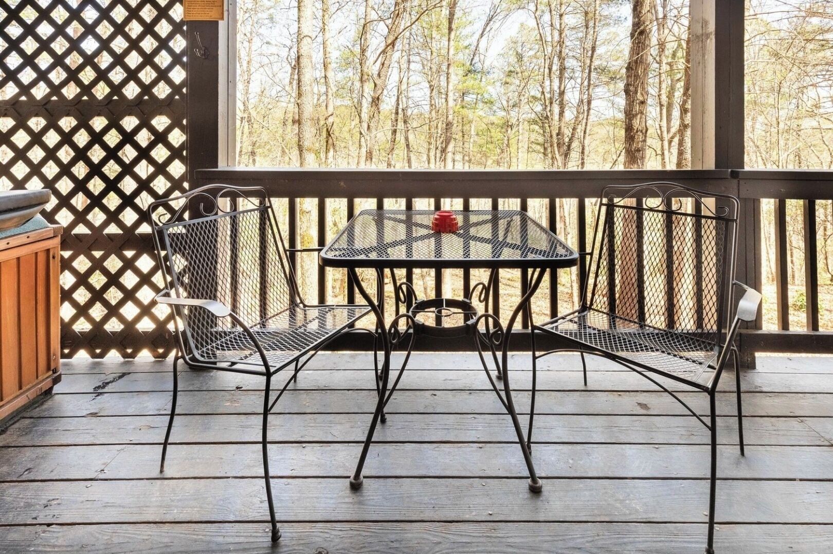 A covered outdoor patio with a metal table and chairs, a wooden cabinet, and a view of trees through a lattice fence.